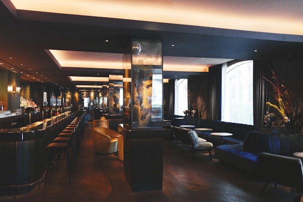 A modern, dimly-lit bar with a sleek, sophisticated interior. It features a long counter with bar stools, and a series of cozy seating areas with plush couches and small round tables. The space is elegantly decorated with dark tones and reflective surfaces, alongside large windows allowing natural light to enter.