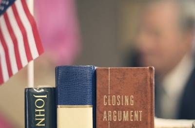 Two books titled 'JOHN...' and 'CLOSING ARGUMENT' are stacked vertically on a table. An American flag is visible to the left of the books. In the background, the blurred figure of a person is seen.