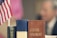 Two books titled 'JOHN...' and 'CLOSING ARGUMENT' are stacked vertically on a table. An American flag is visible to the left of the books. In the background, the blurred figure of a person is seen.