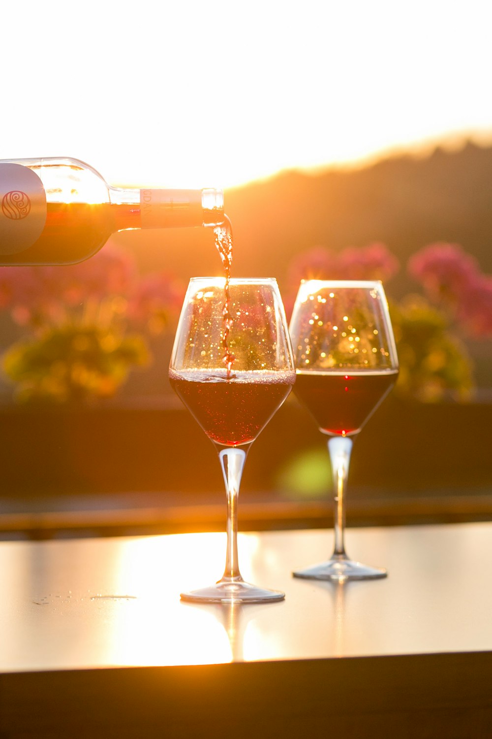 A person pouring red wine into one of two glasses with flowers and the setting sun in the background
