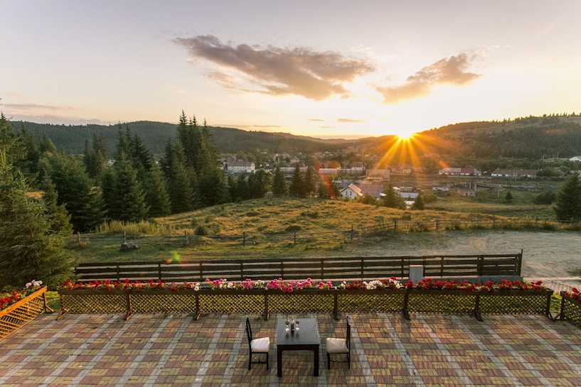 A picturesque landscape featuring a sunset over a small town with pine-covered hills in the background. A foreground patio with a table and chairs is bordered by colorful flowers and overlooks a wide grassy area and a series of homes. The sun's rays spread across the sky, adding warmth and glow to the scene.