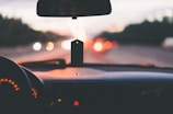 View from inside a car, focusing on the dashboard and a hanging air freshener, with an out-of-focus road and cars in the background. The dashboard's speedometer is partially visible, and the interior is dimly lit, suggesting evening or nighttime. The bokeh effect in the background creates a sense of motion.