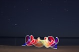 A starry night sky stretches over a calm sea, with the horizon blending into the darkness. In the foreground, a vibrant, abstract light painting features swirling, multicolored ribbons that contrast with the serene background.
