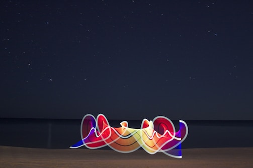 A starry night sky stretches over a calm sea, with the horizon blending into the darkness. In the foreground, a vibrant, abstract light painting features swirling, multicolored ribbons that contrast with the serene background.