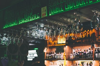A dimly lit bar scene with an overhead rack of hanging wine glasses illuminated by green neon lights. Below, a variety of bottled drinks are arranged on wooden shelves. The ambiance suggests a cozy and atmospheric setting.