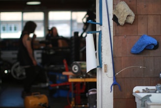 A workshop scene with a focus on a wall holding a roll of paper towels, a blue cloth, and a furry beige item. The background is a garage setting with two blurred figures and various equipment, including an air compressor and tools.