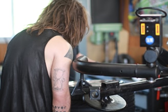 A person with long hair and tattoos on their arm is working in a garage or workshop setting. They are focused on operating machinery or equipment, with a tool or control panel visible in the foreground. The environment has an industrial feel, with blurred details in the background suggesting tools or mechanical elements.