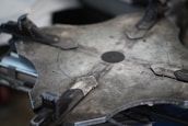 A close-up of a large, industrial-looking metal object with a circular pattern and clamps. The surface appears to be flat and made of a grayish metal with visible textures and markings. Various mechanical components such as clamps are attached around the perimeter, indicating a functional or assembly purpose.