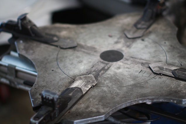 A close-up of a large, industrial-looking metal object with a circular pattern and clamps. The surface appears to be flat and made of a grayish metal with visible textures and markings. Various mechanical components such as clamps are attached around the perimeter, indicating a functional or assembly purpose.