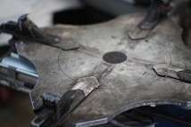 A close-up of a large, industrial-looking metal object with a circular pattern and clamps. The surface appears to be flat and made of a grayish metal with visible textures and markings. Various mechanical components such as clamps are attached around the perimeter, indicating a functional or assembly purpose.
