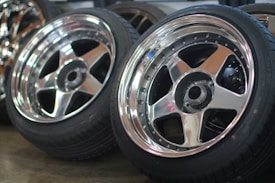 A close-up view of two polished chrome alloy wheels with black rubber tires leaning against each other on a concrete floor. The wheels have a five-spoke design, and their reflective surfaces catch the light, creating a shiny appearance. The background shows a blurred view of additional wheels and a workshop setting.