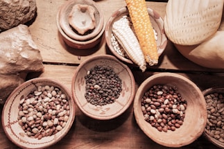 Various types of beans are displayed in clay bowls on a wooden surface alongside different corn cobs. The arrangement includes diverse colors and textures, contributing to an organic and rustic setting.