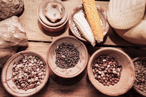 Various types of beans are displayed in clay bowls on a wooden surface alongside different corn cobs. The arrangement includes diverse colors and textures, contributing to an organic and rustic setting.