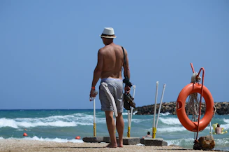 A person stands on a sandy beach facing the ocean waves. The individual is shirtless, wearing a hat and shorts, and holding a pair of shoes in one hand and a plastic bottle in the other. A bright orange lifebuoy is mounted on a nearby stand, and people can be seen swimming in the water in the background. The setting appears calm under a clear blue sky.