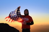 A person stands on a mountain, holding an American flag against a backdrop of a vibrant sunrise or sunset. The individual is wearing a red t-shirt with graphics and a hoodie underneath. The sky transitions from orange near the horizon to a clear blue above.