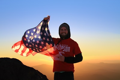 A person stands on a mountain, holding an American flag against a backdrop of a vibrant sunrise or sunset. The individual is wearing a red t-shirt with graphics and a hoodie underneath. The sky transitions from orange near the horizon to a clear blue above.