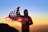 A person stands on a mountain, holding an American flag against a backdrop of a vibrant sunrise or sunset. The individual is wearing a red t-shirt with graphics and a hoodie underneath. The sky transitions from orange near the horizon to a clear blue above.