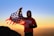 A person stands on a mountain, holding an American flag against a backdrop of a vibrant sunrise or sunset. The individual is wearing a red t-shirt with graphics and a hoodie underneath. The sky transitions from orange near the horizon to a clear blue above.