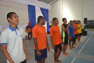 A group of six men, some holding microphones, stand in a line inside a hall. They are casually dressed in colorful t-shirts and shorts. Another person in the background plays a guitar. The setting appears to be a performance or rehearsal space, with blue and white drapery and a speaker visible.