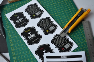 Several printed cocktail labels with a black and white color scheme are placed on a green cutting mat. A yellow utility knife and a metallic ruler are positioned alongside the labels, suggesting a workbench setting. Each label features decorative text and a barcode, with some having additional handwritten colored details.