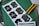 Several printed cocktail labels with a black and white color scheme are placed on a green cutting mat. A yellow utility knife and a metallic ruler are positioned alongside the labels, suggesting a workbench setting. Each label features decorative text and a barcode, with some having additional handwritten colored details.