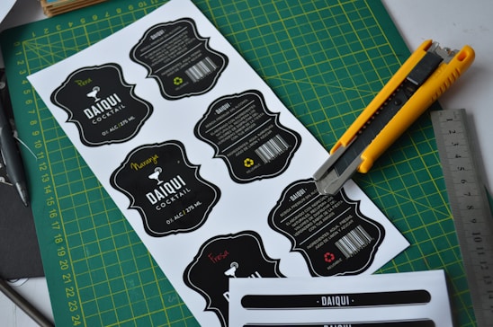 Several printed cocktail labels with a black and white color scheme are placed on a green cutting mat. A yellow utility knife and a metallic ruler are positioned alongside the labels, suggesting a workbench setting. Each label features decorative text and a barcode, with some having additional handwritten colored details.