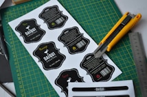 Several printed cocktail labels with a black and white color scheme are placed on a green cutting mat. A yellow utility knife and a metallic ruler are positioned alongside the labels, suggesting a workbench setting. Each label features decorative text and a barcode, with some having additional handwritten colored details.