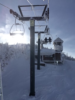 A snowy landscape features a ski lift ascending a slope. The lift towers are covered in snow, and two people are sitting on one of the chairs. In the background, there's a snow-covered cabin and a line of frosted trees under a clear sky with the sun shining from behind a lift tower.