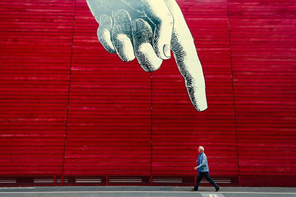 A person walking by a wall painting of a realistic hand with its index finger pointing down.
