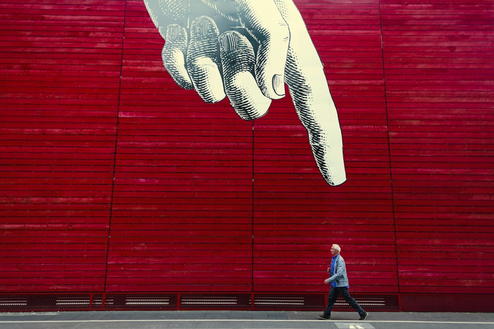 A person walking by a wall painting of a realistic hand with its index finger pointing down.