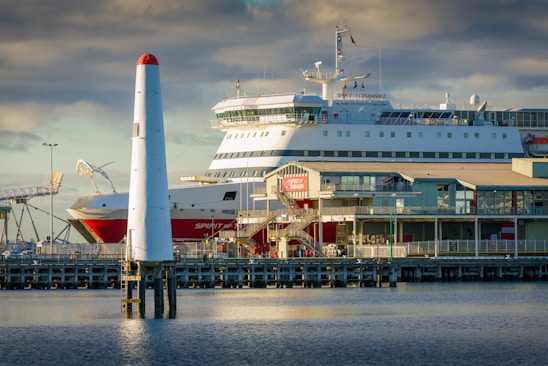 A large ferry is docked at a port with a lighthouse in the foreground. The ferry is white with red accents and several windows, labeled 'Spirit of Tasmania.' There is a clear blue sky with some clouds above, and the sunlight casts a warm glow over the scene.