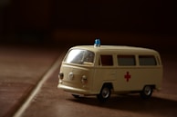 A miniature model of an ambulance with a retro design, featuring a cream color and a red cross symbol on the side. The vehicle has small wheels and a blue emergency light on top. The lighting creates a shadow and an atmospheric effect on the surface it is placed on.