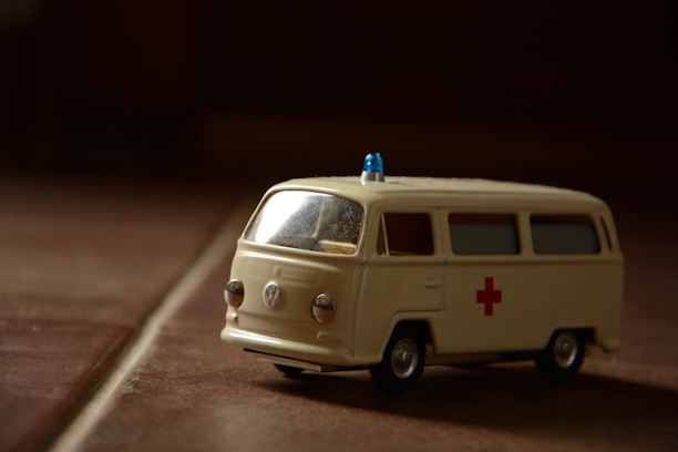 A miniature model of an ambulance with a retro design, featuring a cream color and a red cross symbol on the side. The vehicle has small wheels and a blue emergency light on top. The lighting creates a shadow and an atmospheric effect on the surface it is placed on.