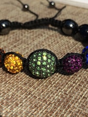 A close-up of a colorful beaded bracelet with round beads encrusted with rhinestones. The beads are strung together with a black cord and feature various colors including yellow, green, purple, and blue. The background consists of a woven fabric texture.