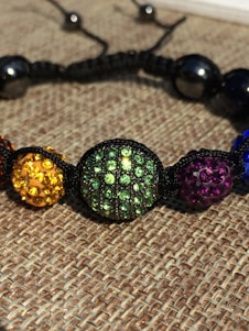 A close-up of a colorful beaded bracelet with round beads encrusted with rhinestones. The beads are strung together with a black cord and feature various colors including yellow, green, purple, and blue. The background consists of a woven fabric texture.