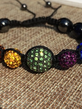 A close-up of a colorful beaded bracelet with round beads encrusted with rhinestones. The beads are strung together with a black cord and feature various colors including yellow, green, purple, and blue. The background consists of a woven fabric texture.