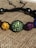 A close-up of a colorful beaded bracelet with round beads encrusted with rhinestones. The beads are strung together with a black cord and feature various colors including yellow, green, purple, and blue. The background consists of a woven fabric texture.