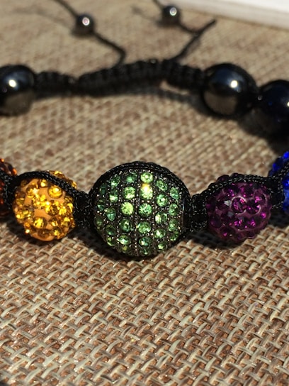 A close-up of a colorful beaded bracelet with round beads encrusted with rhinestones. The beads are strung together with a black cord and feature various colors including yellow, green, purple, and blue. The background consists of a woven fabric texture.