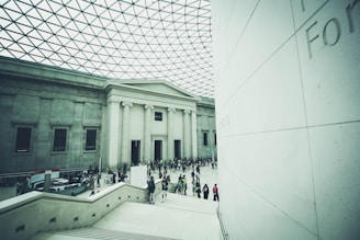 A large indoor area with a high, geometric glass ceiling, resembling a museum or gallery entrance. The space features grand columns and a crowd of people gathered near the entrance, creating a scene of social interaction and exploration.