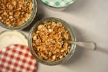 A close-up view of jars filled with a mixture of granola and what appears to be a creamy base. The jars have decorative lids, one with a green checkered pattern and another with a red checkered pattern. A spoon is inserted into one of the jars.