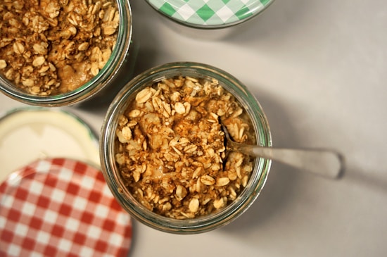 A close-up view of jars filled with a mixture of granola and what appears to be a creamy base. The jars have decorative lids, one with a green checkered pattern and another with a red checkered pattern. A spoon is inserted into one of the jars.