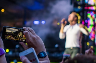 A person holds up a smartphone capturing a live concert performance. The performer on stage is blurred in the background, while the smartphone screen shows them in focus. Concert lights and a vibrant backdrop add to the dynamic atmosphere.
