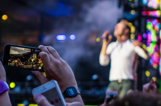 A person holds up a smartphone capturing a live concert performance. The performer on stage is blurred in the background, while the smartphone screen shows them in focus. Concert lights and a vibrant backdrop add to the dynamic atmosphere.