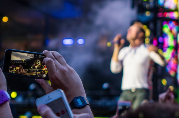 A person holds up a smartphone capturing a live concert performance. The performer on stage is blurred in the background, while the smartphone screen shows them in focus. Concert lights and a vibrant backdrop add to the dynamic atmosphere.