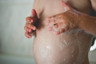 A close-up of a child's torso with hands covered in soap suds gently washing. The lighting is soft, highlighting the texture of the skin and bubbles.