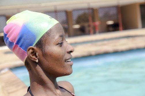 A person wearing a colorful swim cap appears to be relaxing by a swimming pool. Water droplets are visible on the individual's skin, suggesting they have just been swimming. The pool and surrounding area are slightly out of focus in the background.