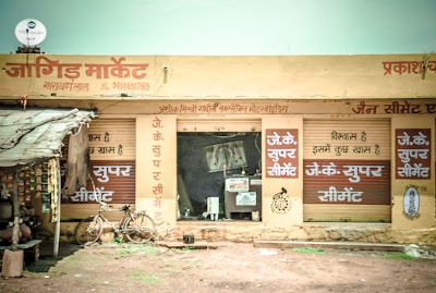 A small market shop in a rural area with signs in Hindi. The shop has a rustic look with handwritten signs advertising 'JK Super Cement'. A bicycle is parked outside the shop, and several packed goods hang on the left. The structure appears to have a corrugated iron roof and natural earthen surroundings.