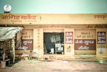 A small market shop in a rural area with signs in Hindi. The shop has a rustic look with handwritten signs advertising 'JK Super Cement'. A bicycle is parked outside the shop, and several packed goods hang on the left. The structure appears to have a corrugated iron roof and natural earthen surroundings.