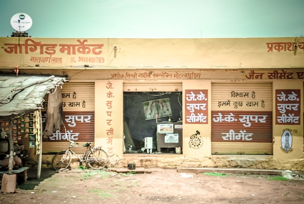A small market shop in a rural area with signs in Hindi. The shop has a rustic look with handwritten signs advertising 'JK Super Cement'. A bicycle is parked outside the shop, and several packed goods hang on the left. The structure appears to have a corrugated iron roof and natural earthen surroundings.