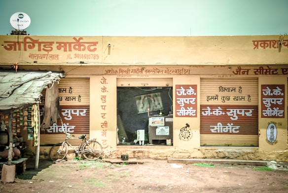 A small market shop in a rural area with signs in Hindi. The shop has a rustic look with handwritten signs advertising 'JK Super Cement'. A bicycle is parked outside the shop, and several packed goods hang on the left. The structure appears to have a corrugated iron roof and natural earthen surroundings.